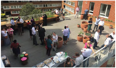 An image of a communal roof terrace. People are stood in groups next to tables of food and there are large decorative plant pots placed around the terrace. 