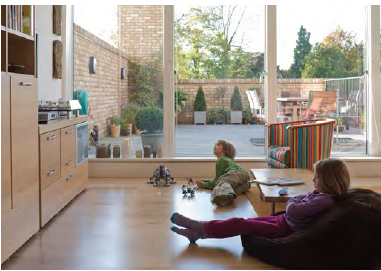 An image showing a private terrace with garden furniture visible through floor to ceiling windows. In the foreground, children are sat in a living room with a beanbag, chair and coffee table.  