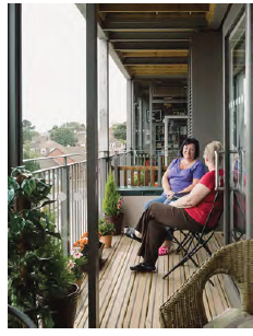 An image of a hanging balcony attached to the side of a residential block of apartments. Two people are shown sat talking on the balcony in garden chairs. Potted plants also line the balcony. 