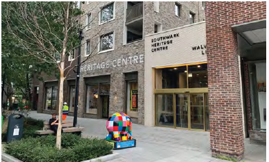 An image of Southwark heritage centre, which features a modern brick façade. There are trees and bushes planted in the pavement in front, along with a colourful art installation. 