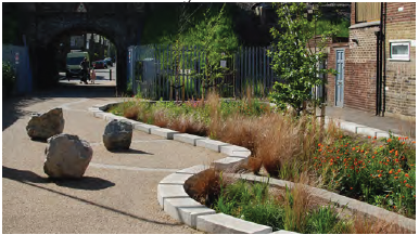An image showing a communal garden with planted beds and integrated play trails. Three large rocks in the pathway create sculptural interest. 